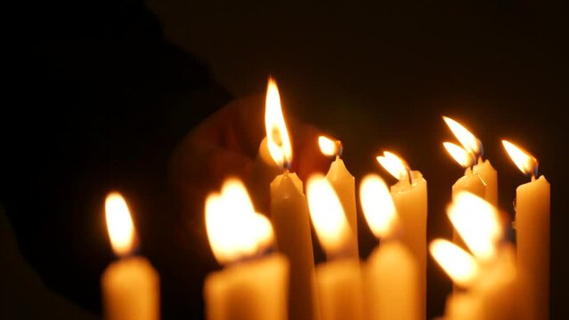Long white religious candles, flames burning at night in the church. A womans hand lights and puts a candle to others