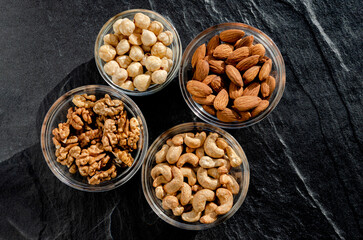 Assortment of nuts in glass bowls on a dark stone table. Cashews, hazelnuts, walnuts, almonds