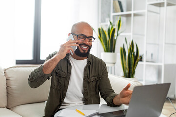 Smiling latin man using laptop and talking on cellphone, communicating with client while working online from home