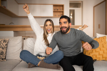 Happy excited young arab man and european woman watching tv, make gesture of victory and success