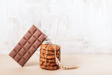A stack of oatmeal chocolate chip cookies tied with twine, a bar of chocolate and a spikelet on a wooden table.