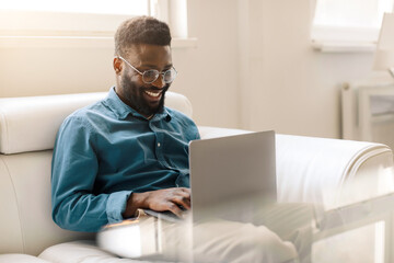 Excited black businessman sitting on sofa in office with laptop, looking at display, taking part in video conference