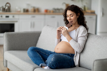 Pregnancy Cravings. Young pregnant woman eating bar of chocolate at home