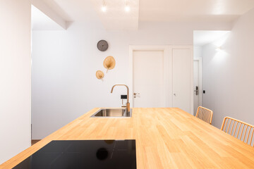 Wooden countertop of kitchen table. Black stove, mixer and sink in chromed metal. The kitchen is lit with bright designer wall lights. Kitchen area at the entrance to studio apartment.