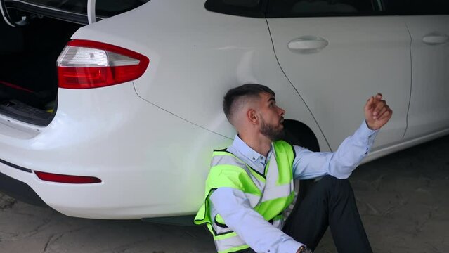 Depressed Young Tired Man In Green Vest Leaning On Car Sitting In Garage Waiting. Portrait Of Stressed Caucasian Guy With Broken Automobile With Flashing Hazard Lights