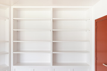 Fototapeta premium Large wooden closed mahogany door with a brass handle for opening. Along the wall is a white open cabinet with shelves for books and interior items for coziness and comfort. The floor is parquet.