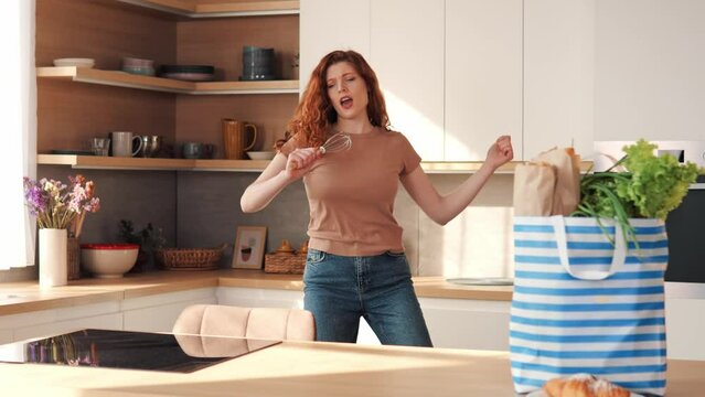 Red hair positive happy woman housewife dancing alone cooking in modern kitchen, funny cheerful active pretty girl preparing food healthy dinner vegetable salad at home having fun listening to music.