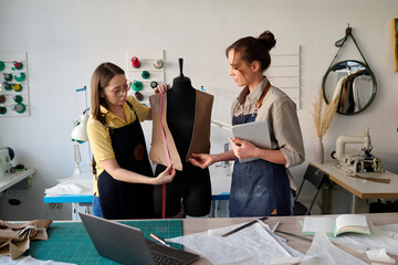 Young subordinate of tailor helping her with taking measures of unfinished leather waistcoat while...