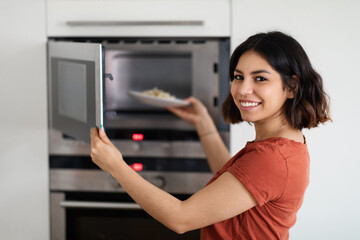 Fototapeta na wymiar Portrait Of Happy Young Arab Woman Using Microwave In Modern Kitchen