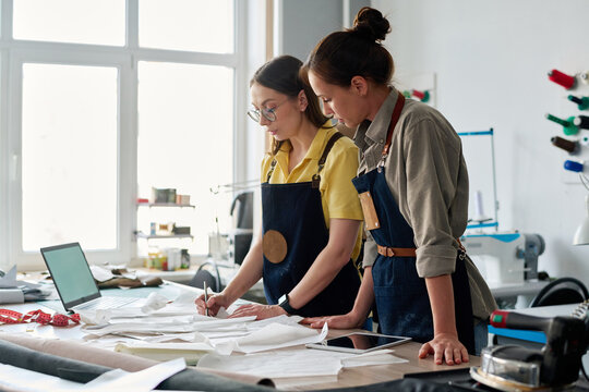 Experienced female tailor putting numbers on sewing paper patterns in atelier while standing by workplace and teaching her subordinate