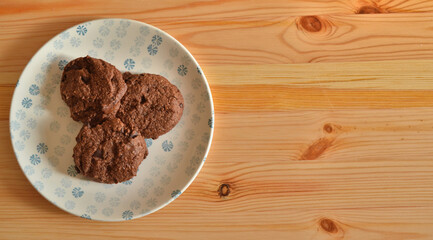 Top view of three dark chocolate cookies with pieces of chocolate on a white and blue porcelain plate on a wooden table.