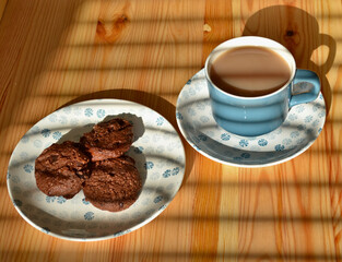 Three dark chocolate cookies with pieces of chocolate on a white and blue porcelain plate, with a blue porcelain cup of coffee with milk on a wooden table on a late afternoon.