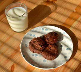 Three dark chocolate cookies with pieces of chocolate on a white and blue porcelain plate, with a glass of milk on a wooden table on a late afternoon.