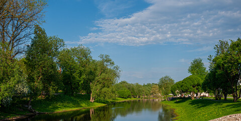 Fototapeta premium Beautiful colorful summer or spring natural landscape with a lake in a park surrounded by green foliage of trees in sunlight and a stone path in the foreground.