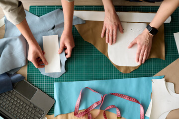 Overview of hands of two female tanners holding sewing paper patterns on pieces of leather while creating new apparel in workshop
