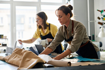 Young female tanner working with sewing patterns and leather textile while her colleague looking through new samples in atelier or studio - obrazy, fototapety, plakaty