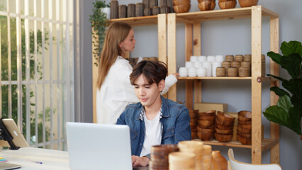 Asian young adult man is telling to Caucasian female coworker to send vase products to him for checking the online purchase order. Two male and female merchants are busy preparing for shipping goods.