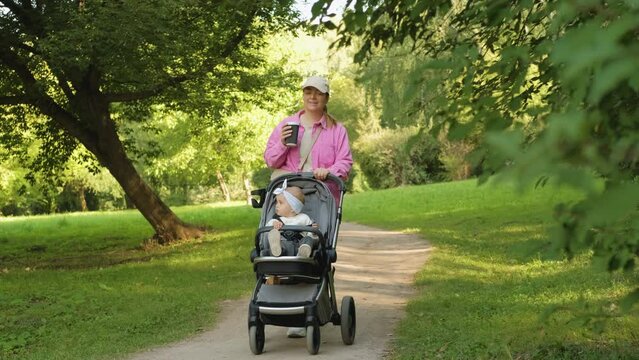 Happy mother drinks coffee while walking in the summer park with her young daughter in a baby carriage outdoors