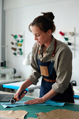 Young female artisan in workwear bending over table in atelier or workshop while using sharp circle...