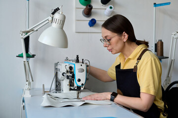 Side view of brunette female tailor or fashion designer bending over electric sewing machine while...