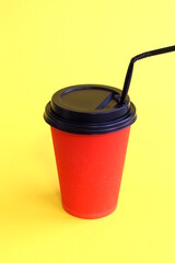 On a yellow background, there is a red glass with a lid and a straw for takeaway hot drinks.	