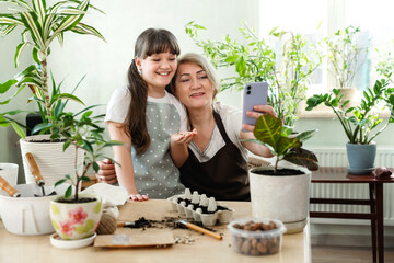 Granddaughter and grandmother are doing selfie on mobile phone in home garden. They are sowing...
