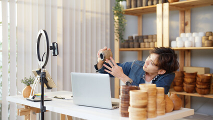 Asian young adult man is giving a presentation about vase product by live streaming online at home. Active male merchant is showing and explaining details of goods to online customers by smartphone.