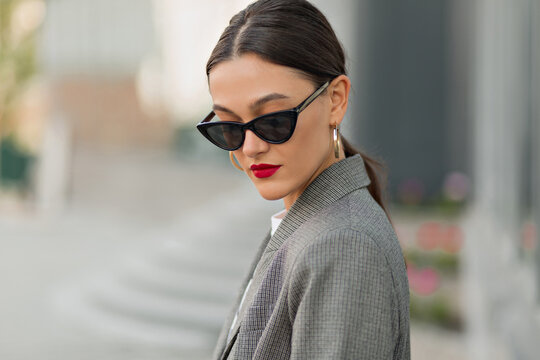 Cheerful adorable lady with dark hair and red lips wearing dark glasses and grey jacket posing on blurred city background. Brunette woman with collected hair. Leisure concept. 