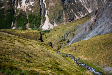 A mountain stream flows down from the rocks with melted snow in the high mountains. Green vegetation in the mountains in summer.