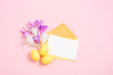 Envelope with blank card, purple crocus flowers, yellow Easter eggs on pink background. Easter greeting card. Top view, flat lay, mockup