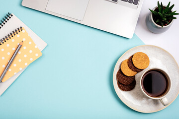 A cup of coffee with cookies, laptop computer, notepads and succulent plant on blue background. Cozy workplace, work from home concept. Top view, flat lay, copy space