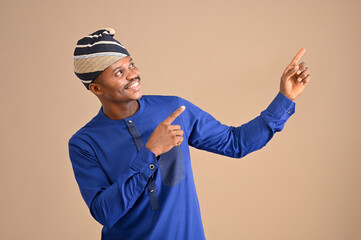 Positive African businessman pointing finger away with toothy smile on his face, empty space for your advertisement, wearing native style attires. Indoor studio shot isolated on brown background.
