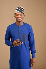 A traditional african man holding his smartphone with his right hand in front of an isolated background his wearing a blue outfits and cap with his smiling at the camera