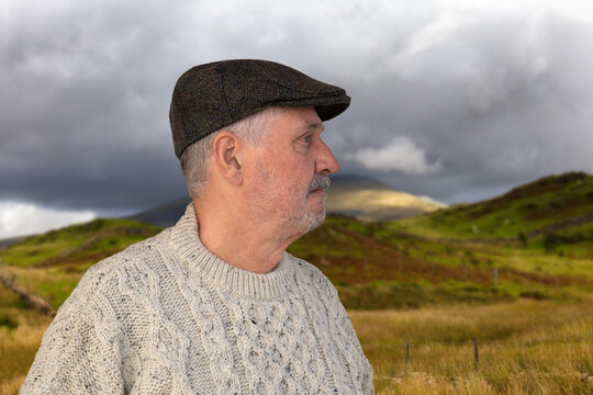 An elderly man wears a stylish Irish sheep's wool sweater and tweet cap. He is standing in a mountainous grassy landscape in Wales.