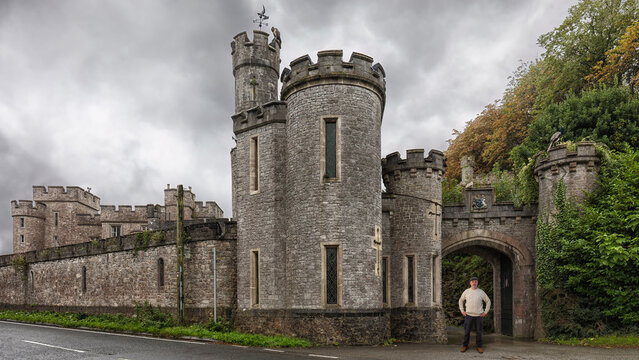 An elderly man stands in a driveway to a British castle. He wears a sheep's wool sweater and a tweet cap. There is a tower on the left.