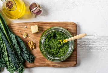 Homemade kale pesto in a glassware with spoon on cutting board over white wooden surface with basic ingredients - kale leaves, walnuts, parmesan, garlic and olive oil