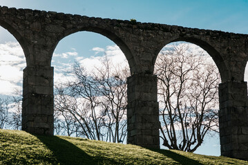 Arches of the Santa Clara Aqueduct that surrounds the church in Vila do Conde, Portugal seen from below, backlit with dry trees, lawn and clouds in the sky