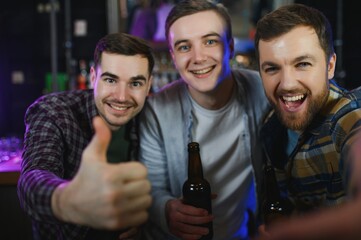 Three young men in casual clothes are smiling, holding bottles of beer while standing near bar counter in pub