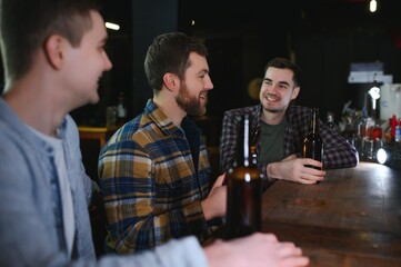 Three young men in casual clothes are smiling, holding bottles of beer while standing near bar counter in pub