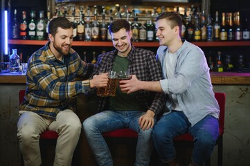 Three young men in casual clothes are smiling, holding bottles of beer while standing near bar counter in pub
