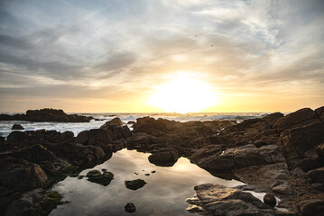 Ball of sun setting among the beach stones and reflecting the clouds in the natural pool. Porto, Portugal