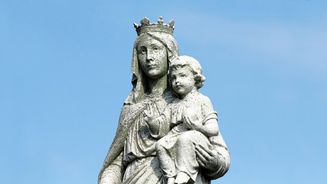 1920s statue of the virgin Mary with baby Jesus in cemetery. Worn, weathered and covered in moss against a blue sky.