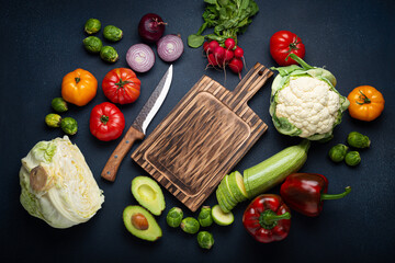 Fresh various vegetables, cut zucchini, wooden cutting board and knife on rustic dark background top view. Cooking vegetarian meal from healthy ingredients, diet food and nutrition concept
