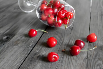 On a wooden table lies a bokkal with cherries, some crumbled .Red cherry berries are scattered on a wooden table. On a wooden board are red cherry berries. Cherries poured out of the glass. - obrazy, fototapety, plakaty