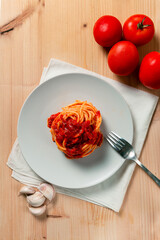 Top vertical view of spaghetti nest with tomato sauce over a wooden table, with a fork, kitchen towel, tomatoes and garlic