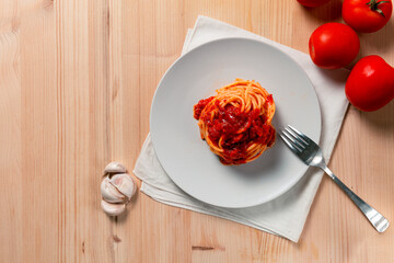 Top view of spaghetti nest with tomato sauce over a wooden table, with a fork, kitchen towel, tomatoes and garlic