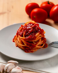Vertical view of spaghetti nest with tomato sauce over a wooden table, with a fork, kitchen towel, tomatoes and garlic