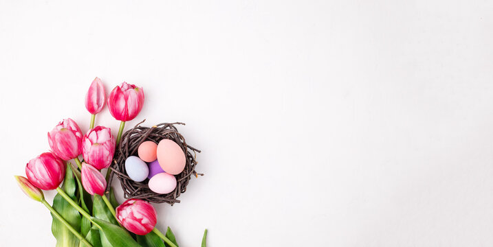 Colorful Easter eggs in decorative nest and tulip flowers on light background, flat lay. Space for text