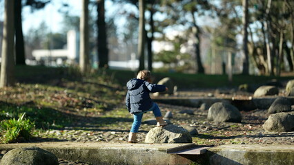 Obraz na płótnie Canvas Carefree child climbing stones and jumping outside in city park. Happy little boy wearing warm jacket and boots. Kid exploring nature