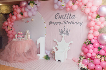 Festive decorations, pink balloons arch, white chair in crown shape, wooden number one. Birthday cake and candy bar. 1 year old girl birthday party photo zone